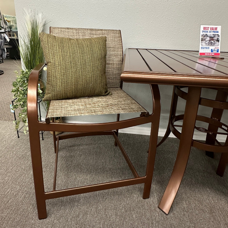 A bronze-finished outdoor dining set consisting of a chair with a woven seat and a cushion, and a matching slatted table, displayed in a showroom.