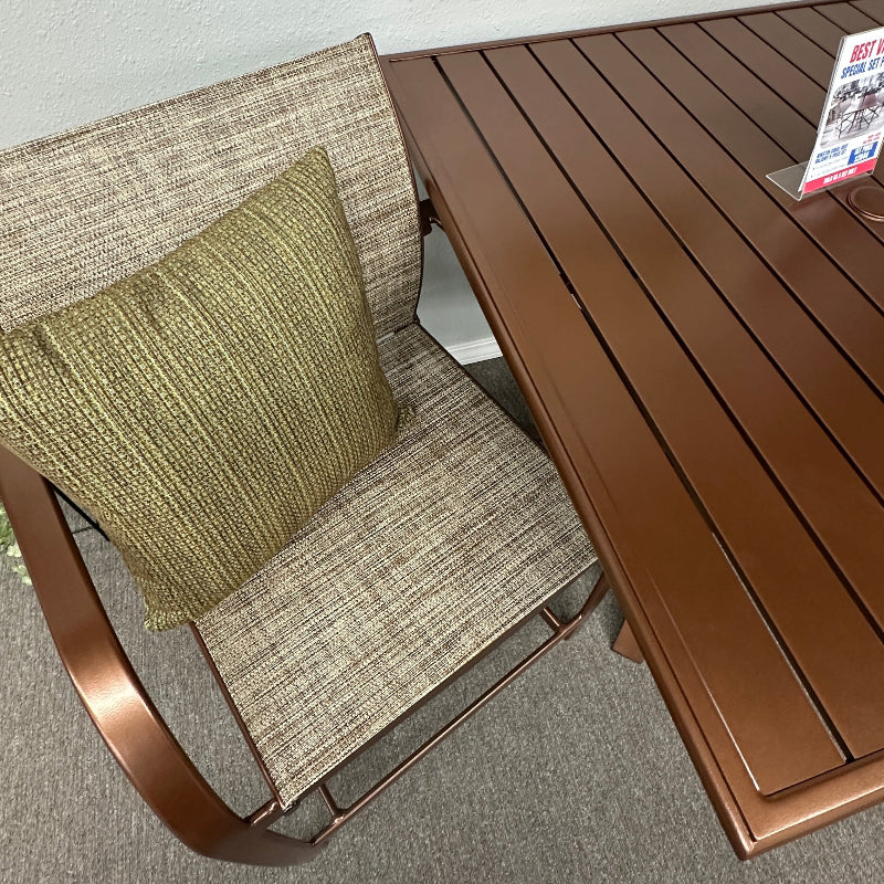 Outdoor patio dining set featuring a brown slatted table and a matching armchair with beige woven fabric upholstery and a green throw pillow, shown on a gray carpet against a white wall.