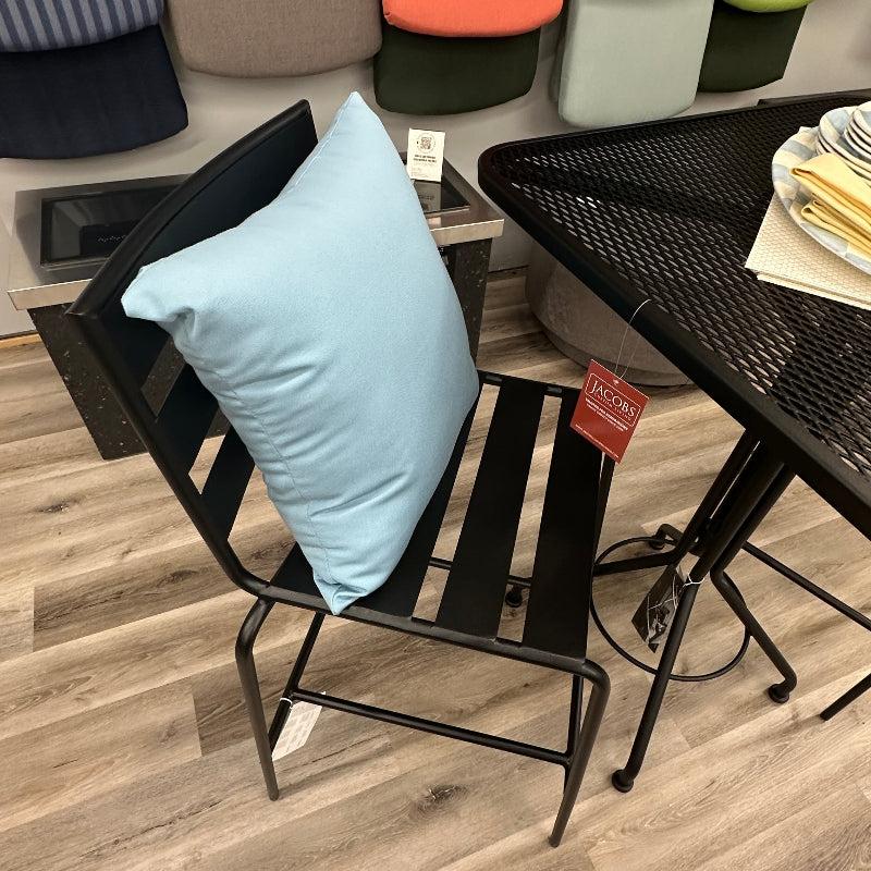 A black metal patio chair with a light blue throw pillow, shown in a furniture store setting with wood-look flooring and a black mesh table nearby.