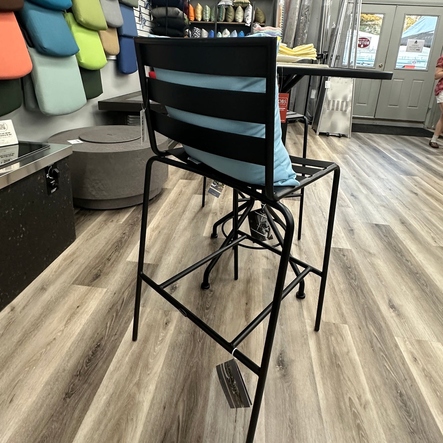 A black metal high bar stool with a light blue cushion on a wood-look floor inside a furniture or upholstery showroom.