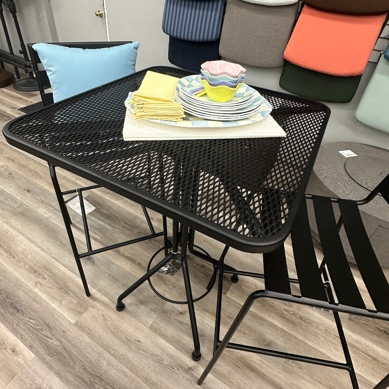 A black metal mesh dining table with matching black chairs in a furniture showroom, styled with a stack of plates and yellow napkins on top. Color swatch displays and gray wood-look flooring are visible in the background.
