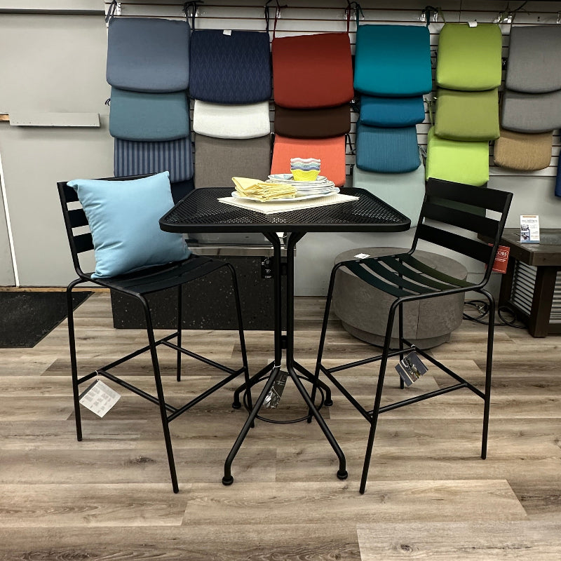 A black metal bar-height dining set consisting of a round table and two matching chairs, displayed in a furniture showroom with a wall of colorful throw pillows and cushions behind it.