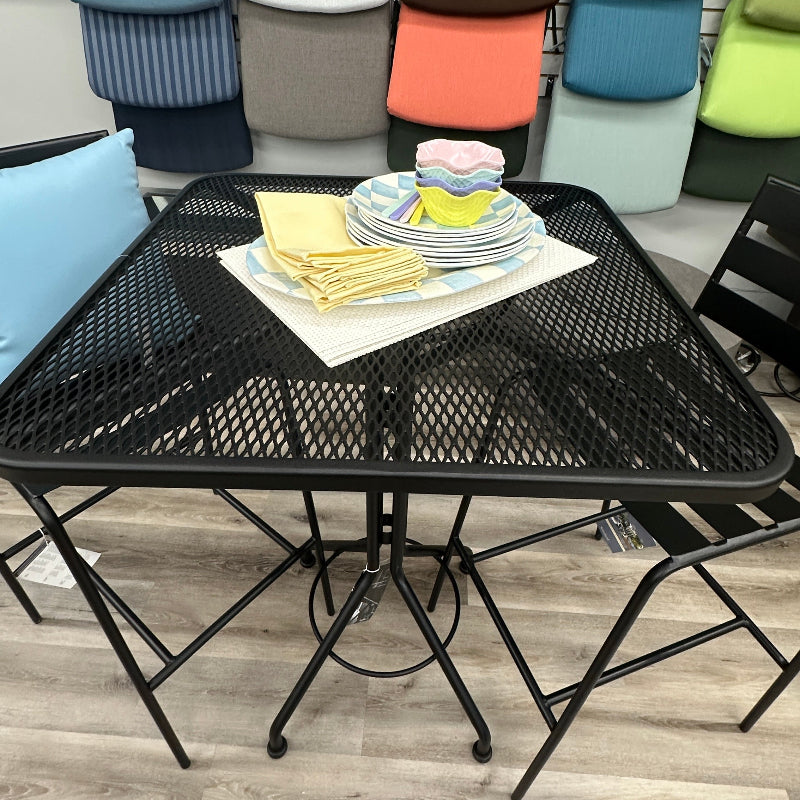 A black metal mesh square dining table with matching black chairs, shown in a furniture store. A stack of folded napkins and plates sits on the table, and a wall behind it displays numerous upholstered chair cushions in various colors and fabrics.