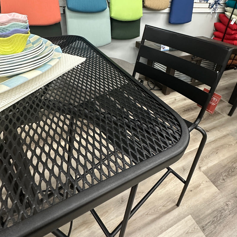 A black metal mesh patio dining table with a matching black metal chair, shown in a furniture store with colorful cushion samples stacked on a wall behind it.