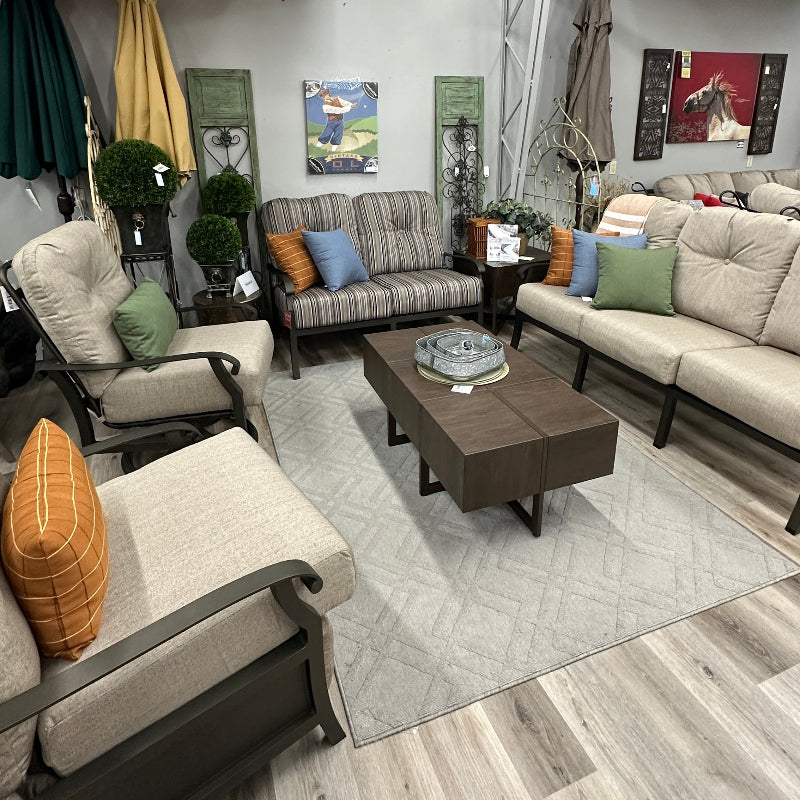 A staged outdoor living room furniture display featuring a striped sofa, a beige sectional sofa, a matching armchair, a dark wood coffee table, and a light gray patterned rug on a wood-look floor.