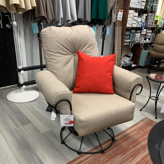 A small outdoor patio swivel rocker chair with a beige fabric cushion and a red accent pillow, featuring a black wrought iron frame, displayed in a retail setting.