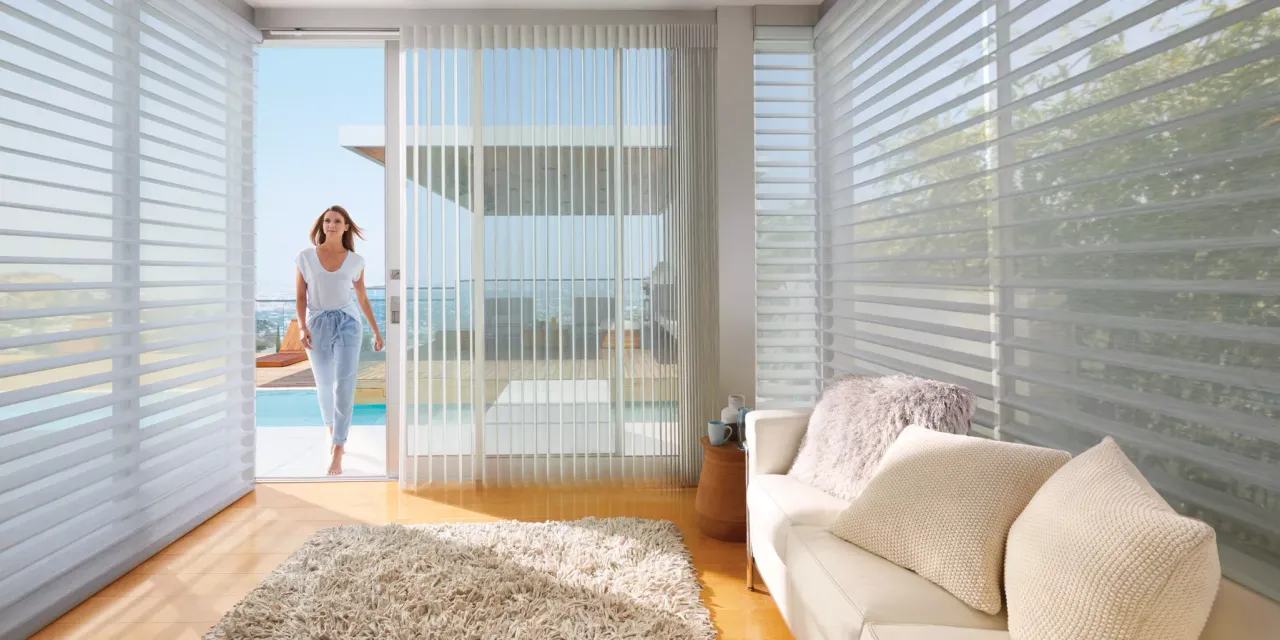 Living room with white window blinds and a woman standing by a glass door.